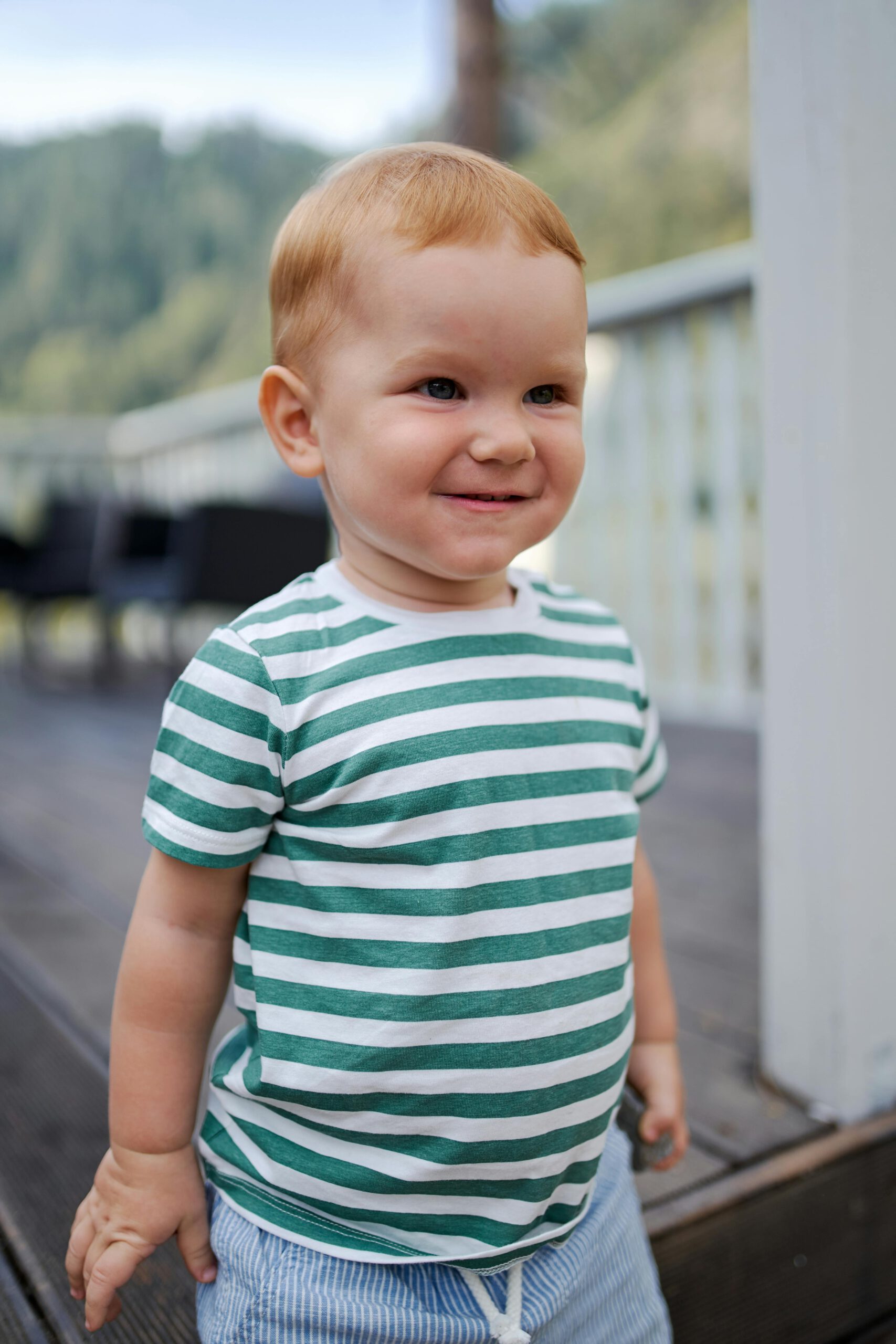 Cute toddler smiling outdoors in green striped shirt on a wooden terrace.